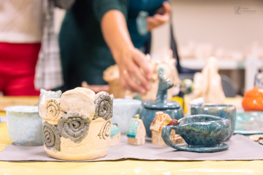 Prace ceramiczne w różnych kolorach ustawione na stole, w tle ręka sięgająca, po któryś z ustawonych elementów.