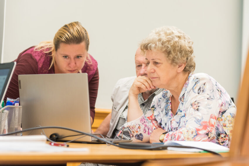 Dwoje Seniorów siedzących przy ławce oraz stojąca obok młodsza pani wpatrzeni w ektan laptopa.