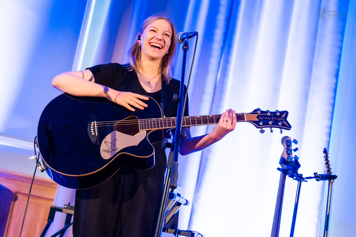 Uśmiechnięta młoda dziewczyna z gitarą w rękach w pomieszczeniu oświetlonym niebieskim światłem.