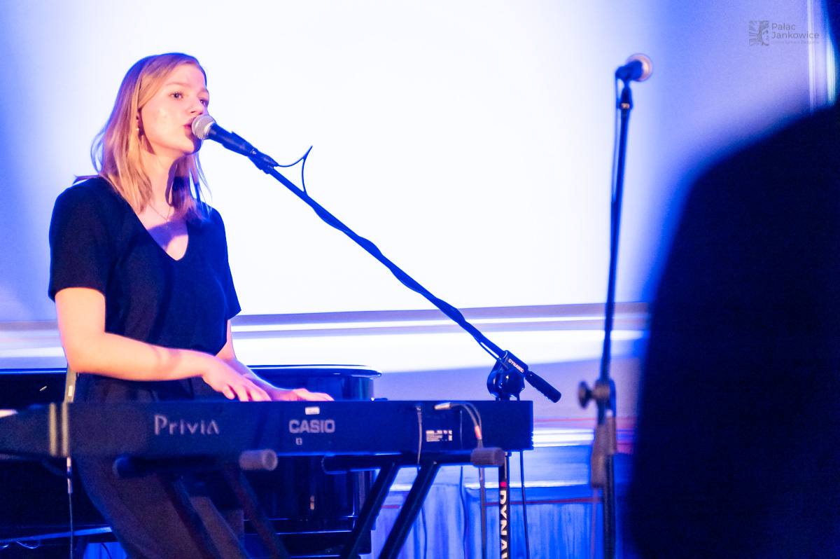Młoda dziewczyna przy pianinie elektrycznym, śpiewająca do mikrofonu na statywie.