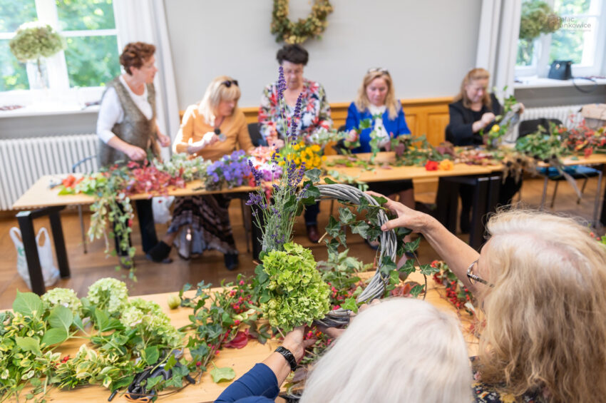 Seniorki tworzą wianki w dużej sali z ławkami, na których rozłożone są kolorowe kwiaty i zielone rośliny.